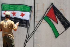 A person facing away from the camera carrying a spray can in front of a wall with the Syrian Independence Flag and the Palestinian Flag spraypainted on it.